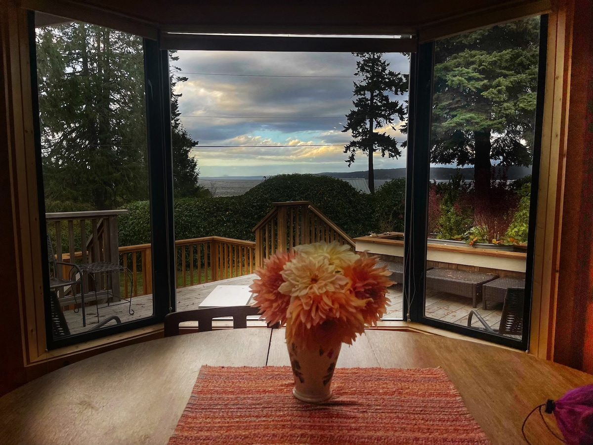A view from a window showcases a serene landscape, featuring water and distant mountains framed by lush greenery. In the foreground, a vase with flowers rests on a wooden table, adding a touch of color to the cozy interior.