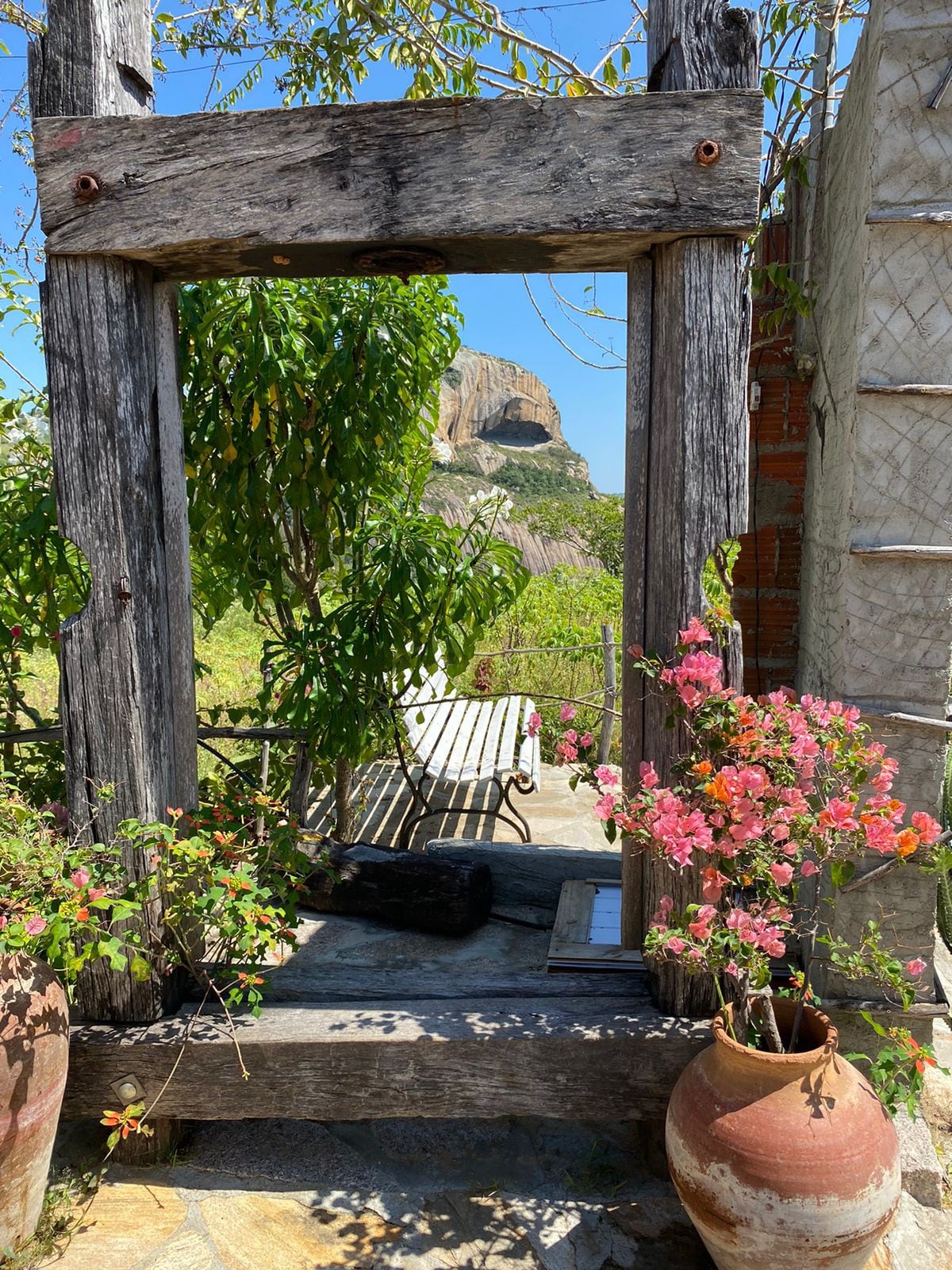 A rustic wooden frame creates a scenic view of the outdoor area, emphasizing colorful flowers and greenery. A stone pathway leads to a bench, with a backdrop showcasing a prominent rocky outcrop under a clear blue sky.