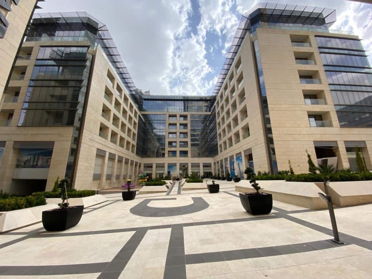 A modern building façade is viewed from the courtyard, showcasing a sleek design with large glass windows and neutral stone exteriors. Potted plants are arranged throughout the courtyard, and a patterned pathway leads the eye toward the entrance, enhancing the urban ambience.
