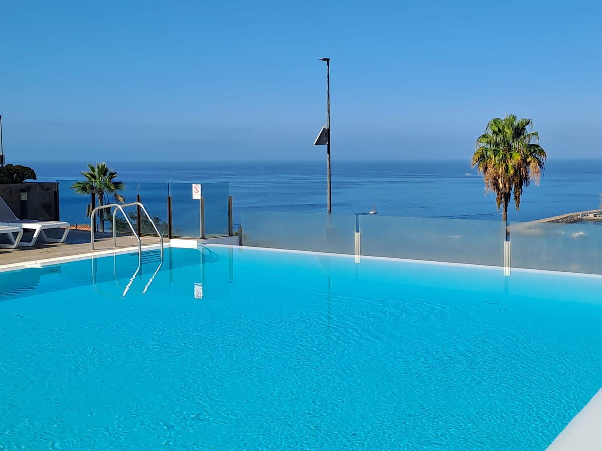 A serene swimming pool is surrounded by clear glass barriers, offering unobstructed views of the Atlantic Ocean. Palm trees are visible nearby, and a blue sky reflects on the water's surface, enhancing the tranquil atmosphere.