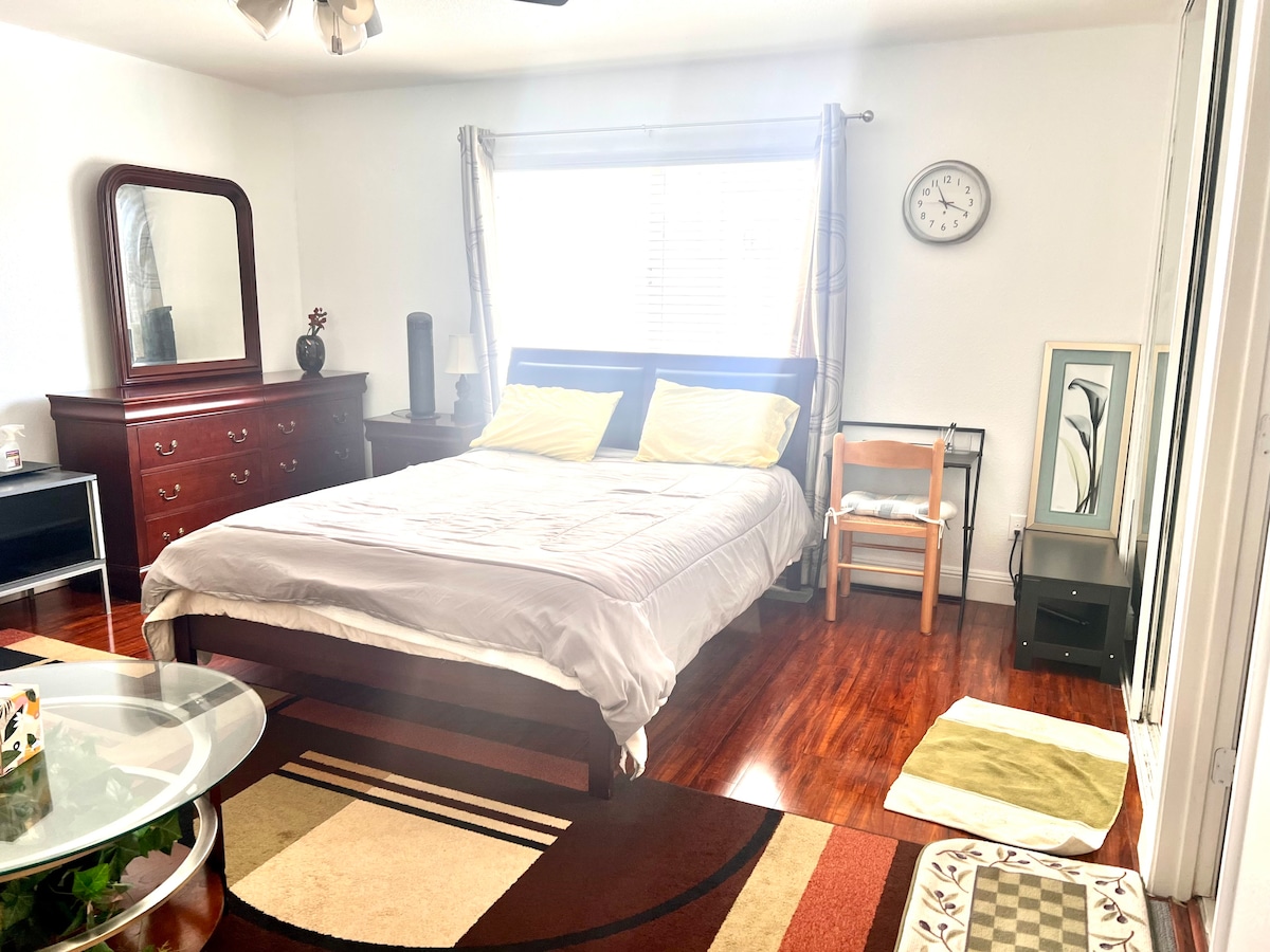 A spacious bedroom features a large bed with yellow pillows. A wooden dresser and a mirror are positioned nearby. A round table adds functionality, while a chair and a small side table complete the layout. Natural light enters through the window, illuminating the warm wooden floor.
