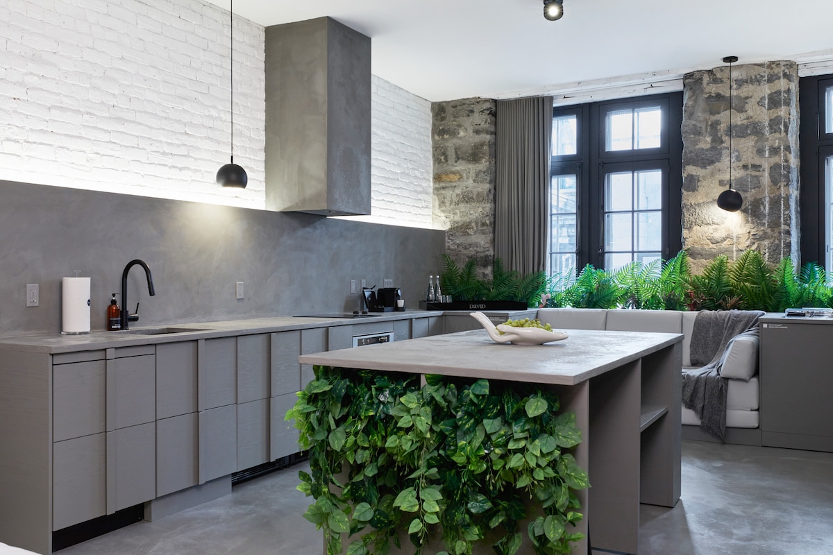 A modern kitchen features sleek gray cabinetry and a spacious island adorned with greenery. The kitchen is illuminated by pendant lighting, and large windows allow natural light to highlight the exposed brick walls. A comfortable seating area is visible in the background.