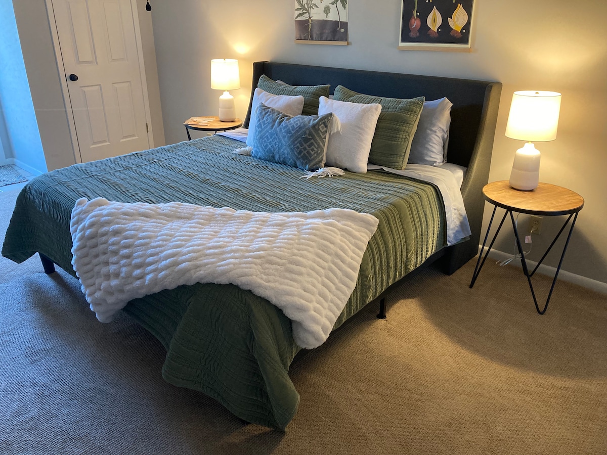 A spacious bed is dressed in muted green bedding, accompanied by a white textured throw. Each side table is topped with a lamp, providing soft illumination. Light enters through a nearby door, enhancing the warm tones of the room.