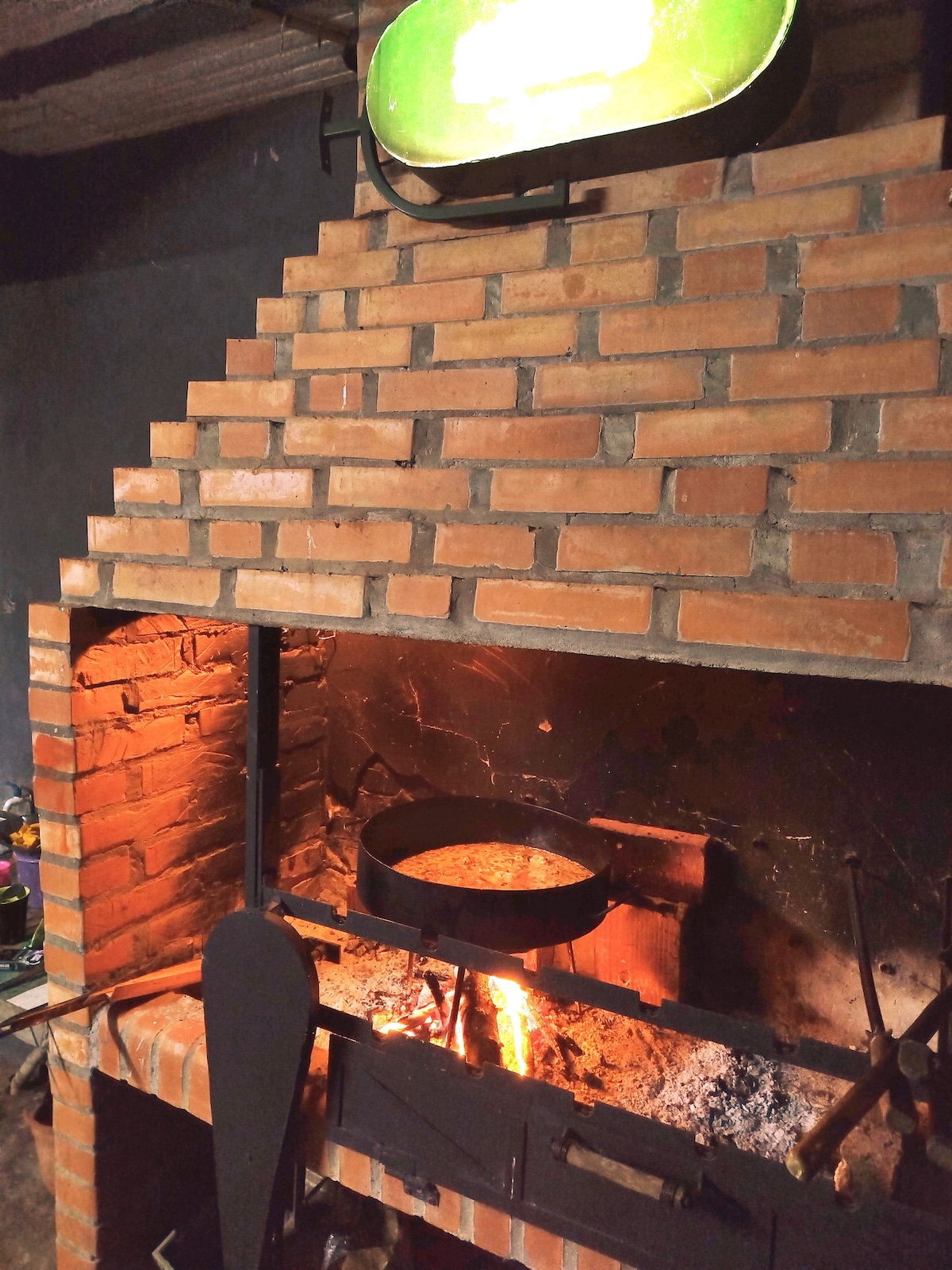 A traditional brick fireplace is featured, with a circular metal pan resting above a vibrant flame. The warm glow from the fire illuminates the surrounding bricks, creating a cozy setting. The fireplace structure includes a sturdy metal stand, enhancing the rustic appeal.
