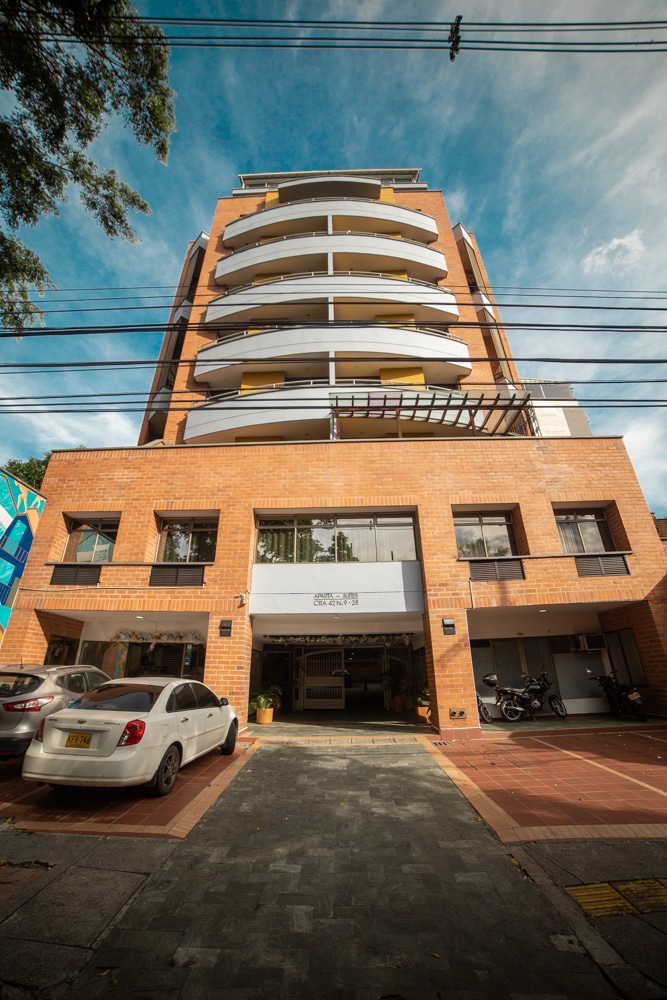 A modern brick building is presented with multiple floors and a series of circular balconies. The entrance is accessed through a spacious lobby area visible through large glass windows, framed by parked cars and well-maintained landscaping.
