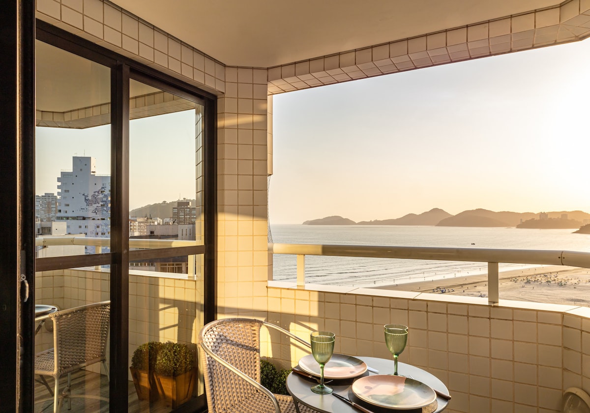 A balcony is shown with a view overlooking the beach and mountains in the distance. Two chairs are positioned around a small table, set with two glasses. The warm glow of sunset illuminates the scene, enhancing the tranquil atmosphere.