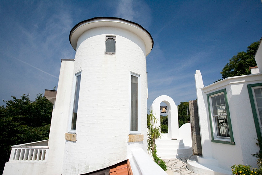 A white-walled structure features a round tower, complementing the modern architectural design. Sunlight brightens the exterior, showcasing arched doorways and lush greenery in the vicinity. A smooth stone pathway leads towards an inviting entrance.