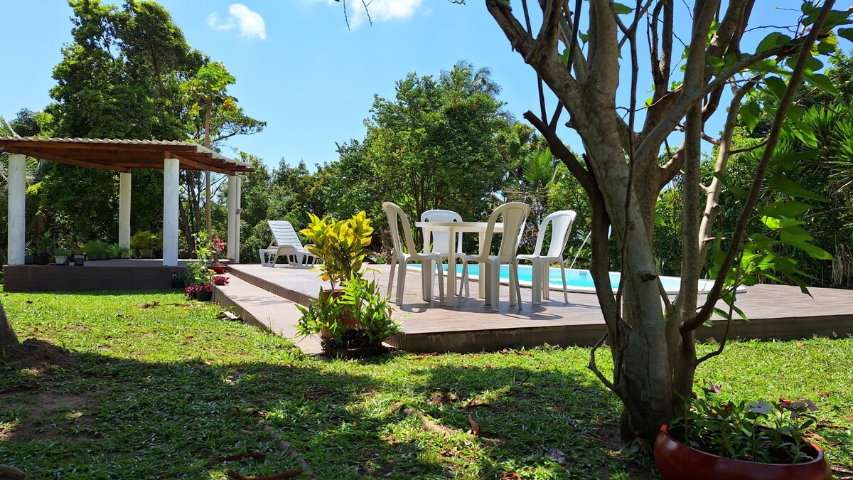 A serene outdoor area is depicted, featuring a wooden deck with a dining table and chairs. A swimming pool is visible in the background, surrounded by lush greenery and flowerbeds. Sun loungers are positioned alongside the pool, providing spaces for relaxation.