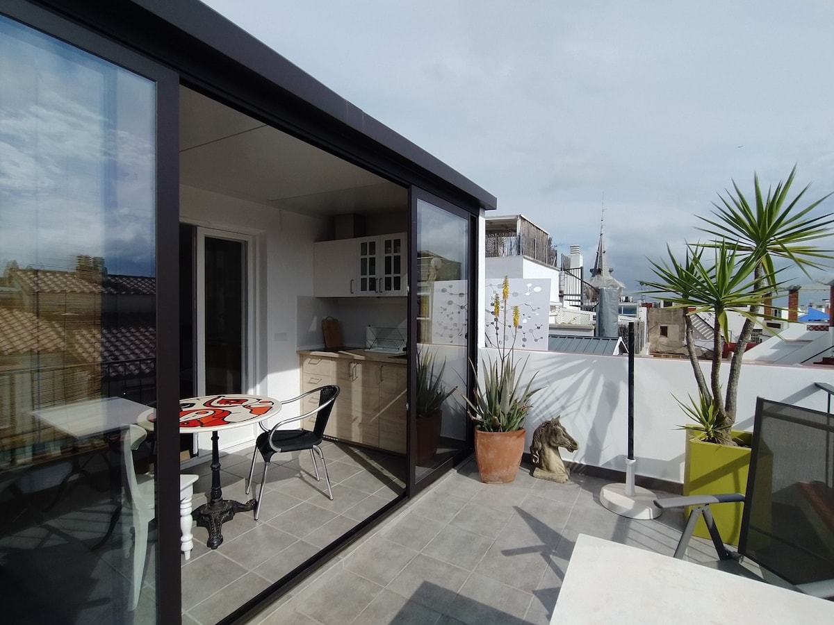 A spacious terrace presents outdoor dining with a table set for two. Glass sliding doors provide access to a kitchenette area, while potted plants add greenery to the space. The views of the surrounding rooftops and distant spires are visible in the background.
