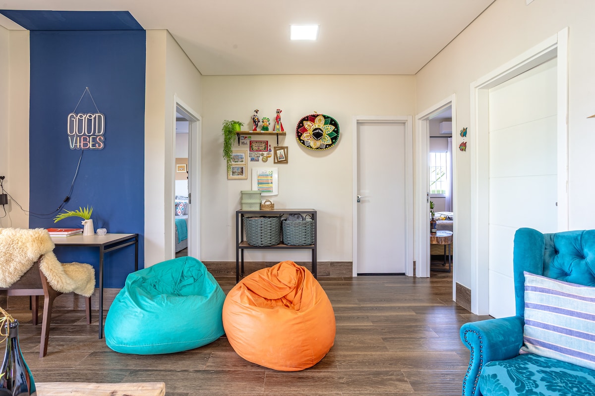 A relaxed sitting area features two bean bag chairs in vibrant teal and orange. Light wood flooring complements the soft, neutral wall tones. Decorative items adorn the walls, with a glimpse of the adjoining rooms visible through open doors.