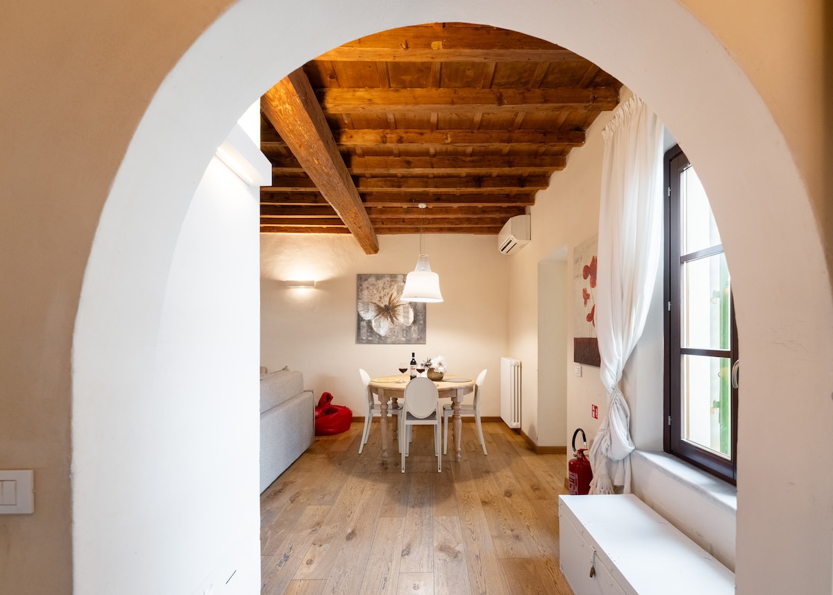 An arched entrance leads into a bright, spacious living area featuring a wooden ceiling and light oak flooring. A dining table with white chairs is visible, complemented by a cozy sofa. Natural light filters in through large windows adorned with sheer curtains.