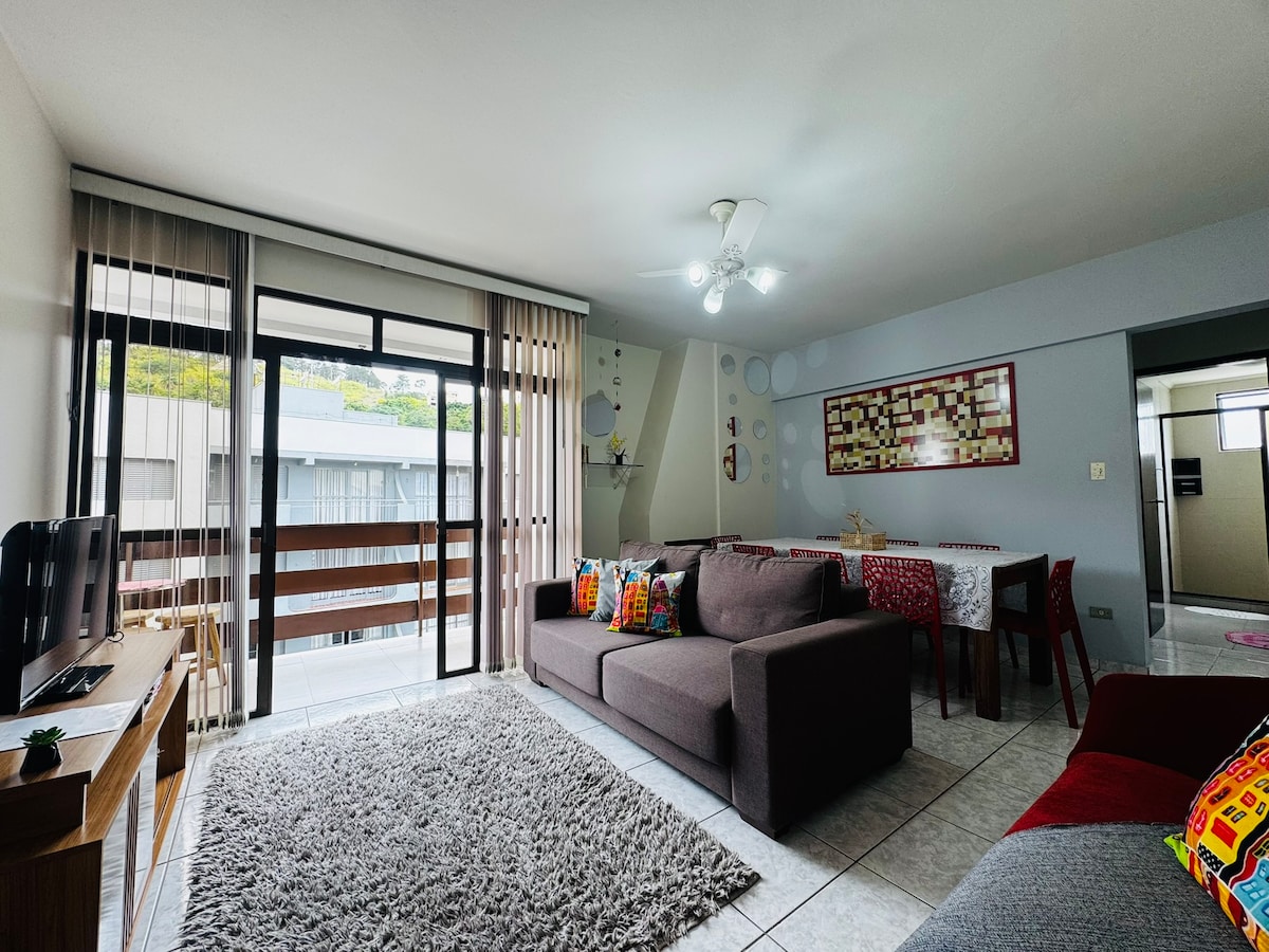 A spacious living area is showcased, featuring a comfortable seating arrangement with colorful throw pillows. A wooden dining table is visible, accompanied by light streaming through large glass doors that open to a balcony. Soft carpeting adds warmth to the tiled floor.