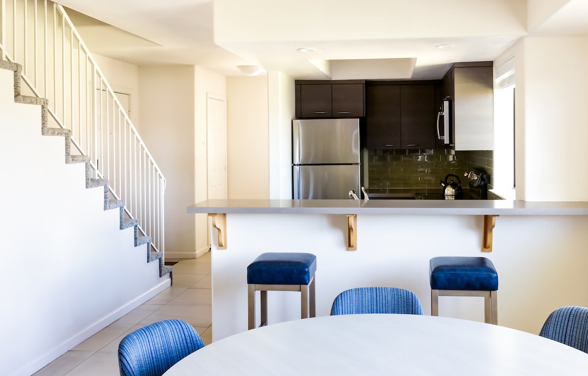 A modern kitchen area is visible with stainless steel appliances and dark cabinetry. The white countertop offers seating with blue bar stools. A round dining table is positioned nearby, surrounded by striped chairs. An open staircase leads to the upper level, enhancing the spacious feel.