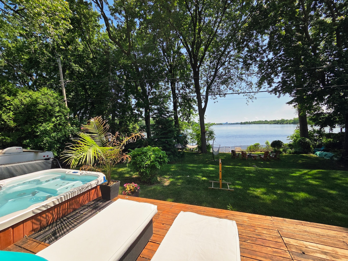 An outdoor deck features a hot tub and two lounge chairs, surrounded by lush greenery. A view of the river is visible beyond the landscaped yard, where a seating area has been arranged for relaxation. The scene is set under a clear blue sky.