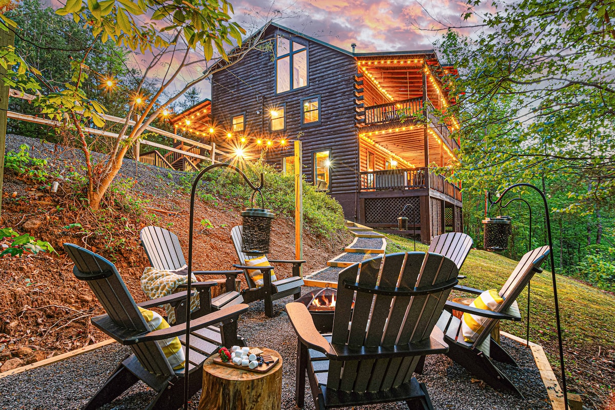A charming cabin exterior is showcased, surrounded by trees and enhanced by warm string lights. Several Adirondack chairs are arranged around a fire pit, inviting gatherings, while a winding path leads to the cabin's multiple decks and outdoor living spaces.