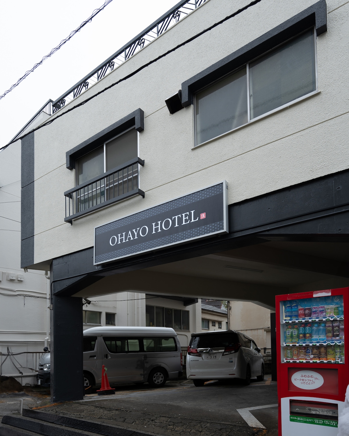 Exterior of Ohayo Hotel with a sign above a parking area and a red vending machine in the foreground.