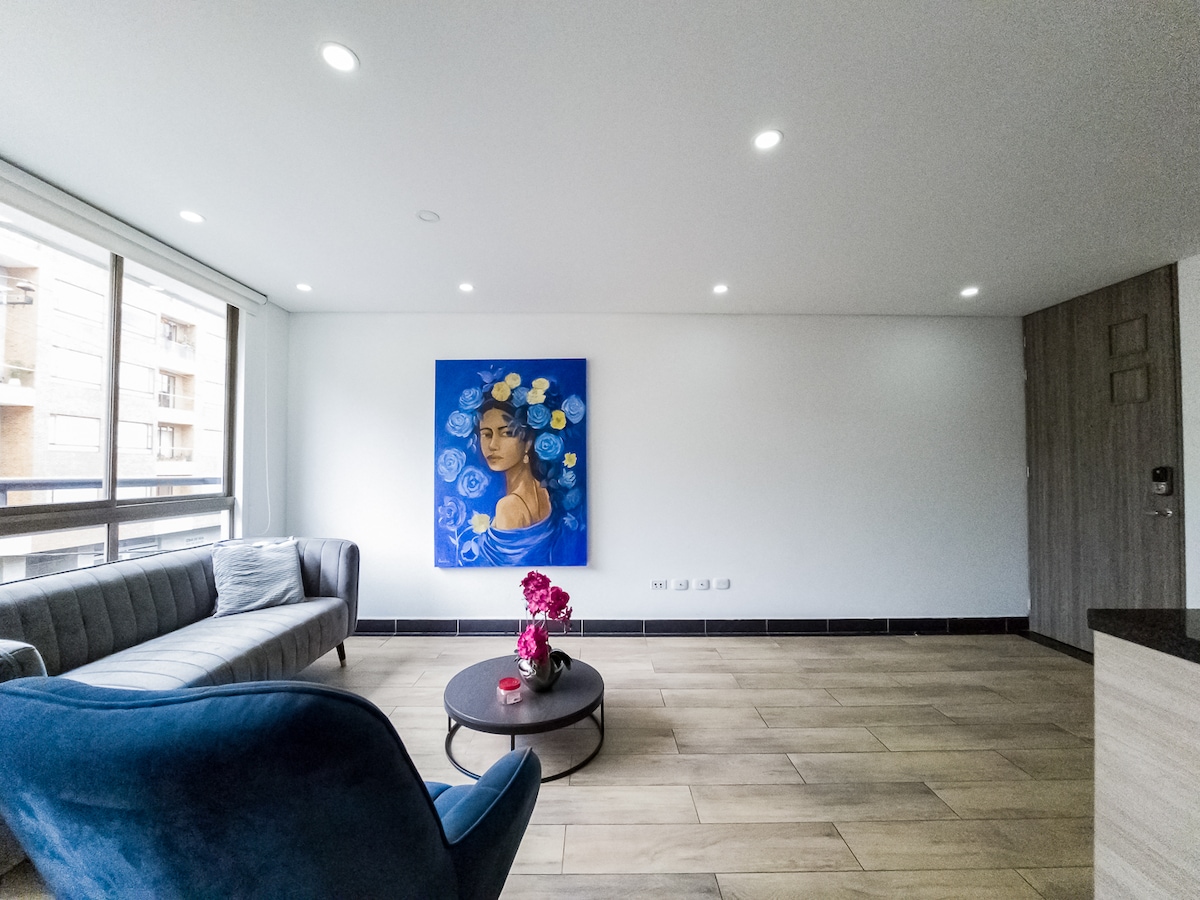 A modern living area features a gray sofa and a blue armchair, complemented by a round coffee table with a floral arrangement. A vibrant blue artwork is mounted on a white wall, enhancing the bright and open feel of the space, which is illuminated by recessed ceiling lights.