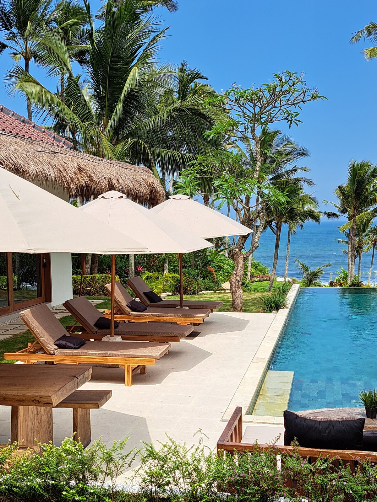A serene outdoor space is presented, featuring a sparkling pool surrounded by lush greenery. Sun loungers with umbrellas are positioned beside the pool, offering a relaxing area to enjoy the ocean view. A dining table is set nearby for al fresco meals.