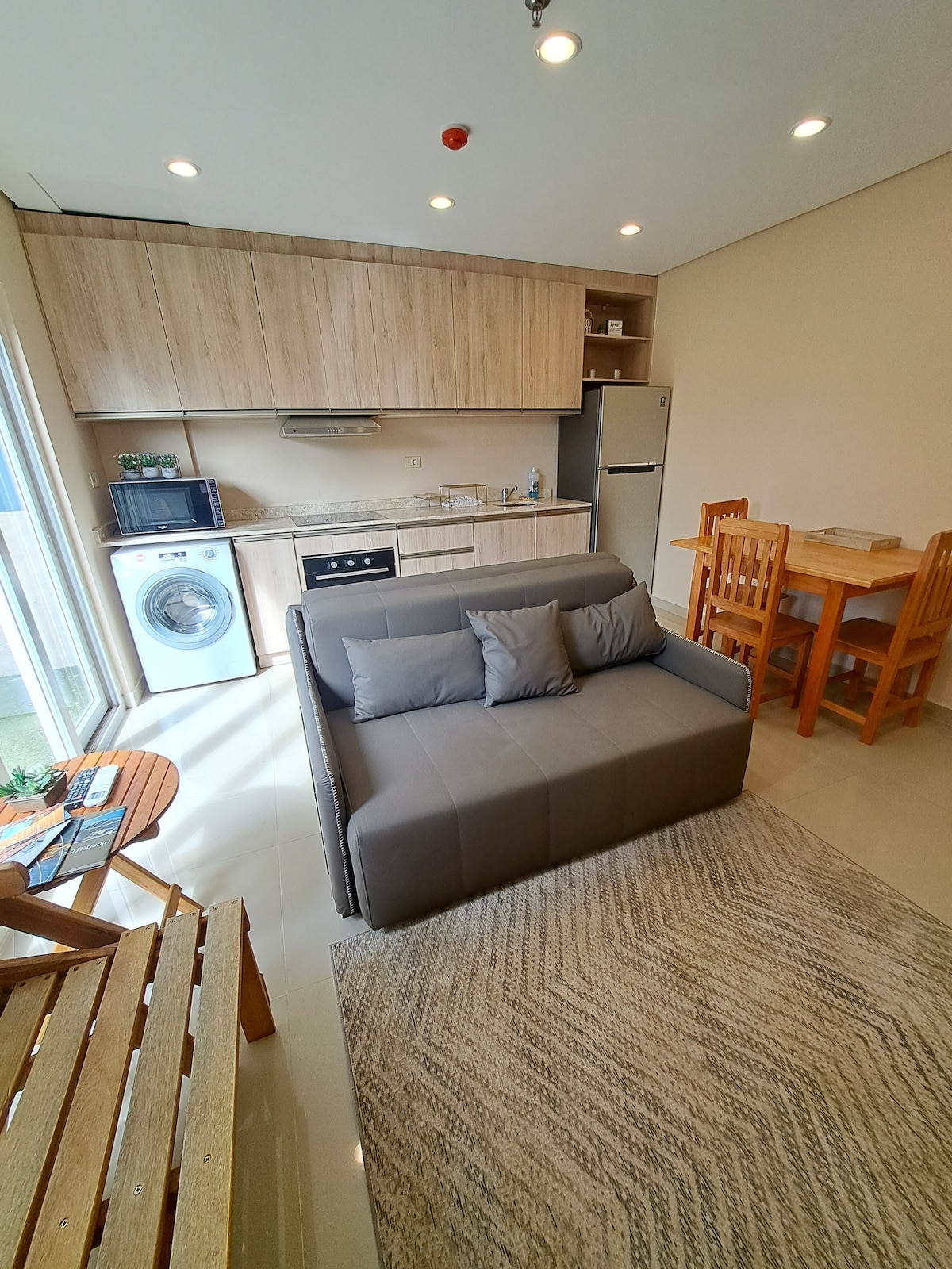 A modern kitchen area features light wood cabinetry and a dual-purpose living space with a gray sofa. A dining table with four wooden chairs is positioned beside the kitchen. Natural light enters through large glass doors leading to a balcony.