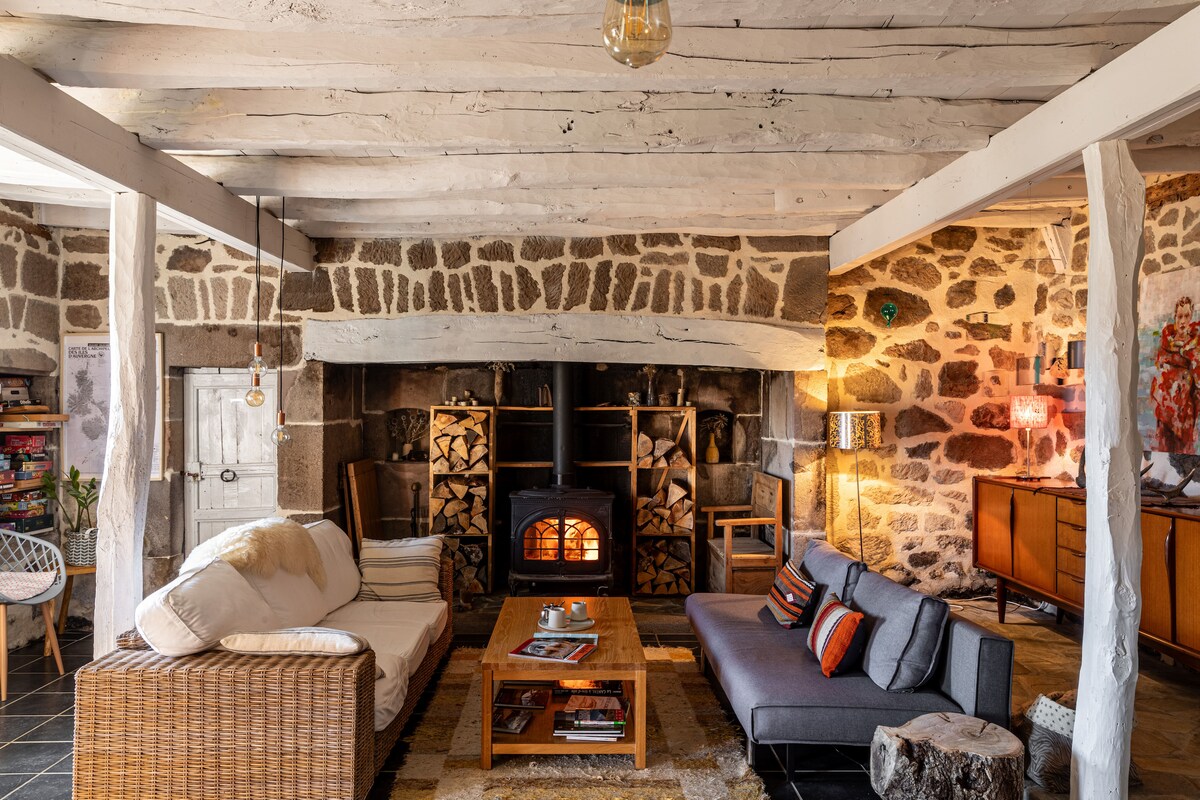 A spacious living area features textured stone walls and traditional wooden beams. A cozy seating arrangement consists of two sofas and a coffee table, with a wood-burning stove providing warmth. Shelves display books, while soft lighting enhances the inviting atmosphere.