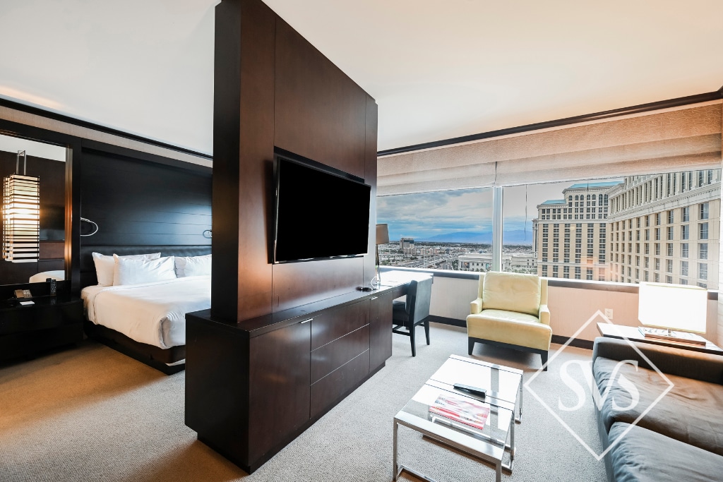 The studio suite features a central room divider with a flat-screen television. A spacious bed is visible, along with a seating area that includes a light-colored armchair and a coffee table. Large windows provide a view of the Las Vegas skyline.