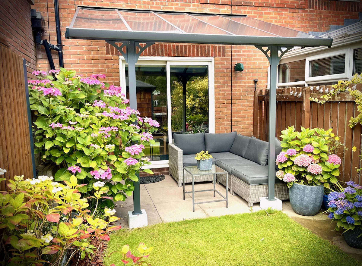 A shaded seating area is surrounded by blooming hydrangeas and lush greenery. A cozy outdoor sofa is accompanied by a small table, inviting relaxation. The patio leads to a glass door opening into the home, creating a seamless connection between indoor and outdoor spaces.