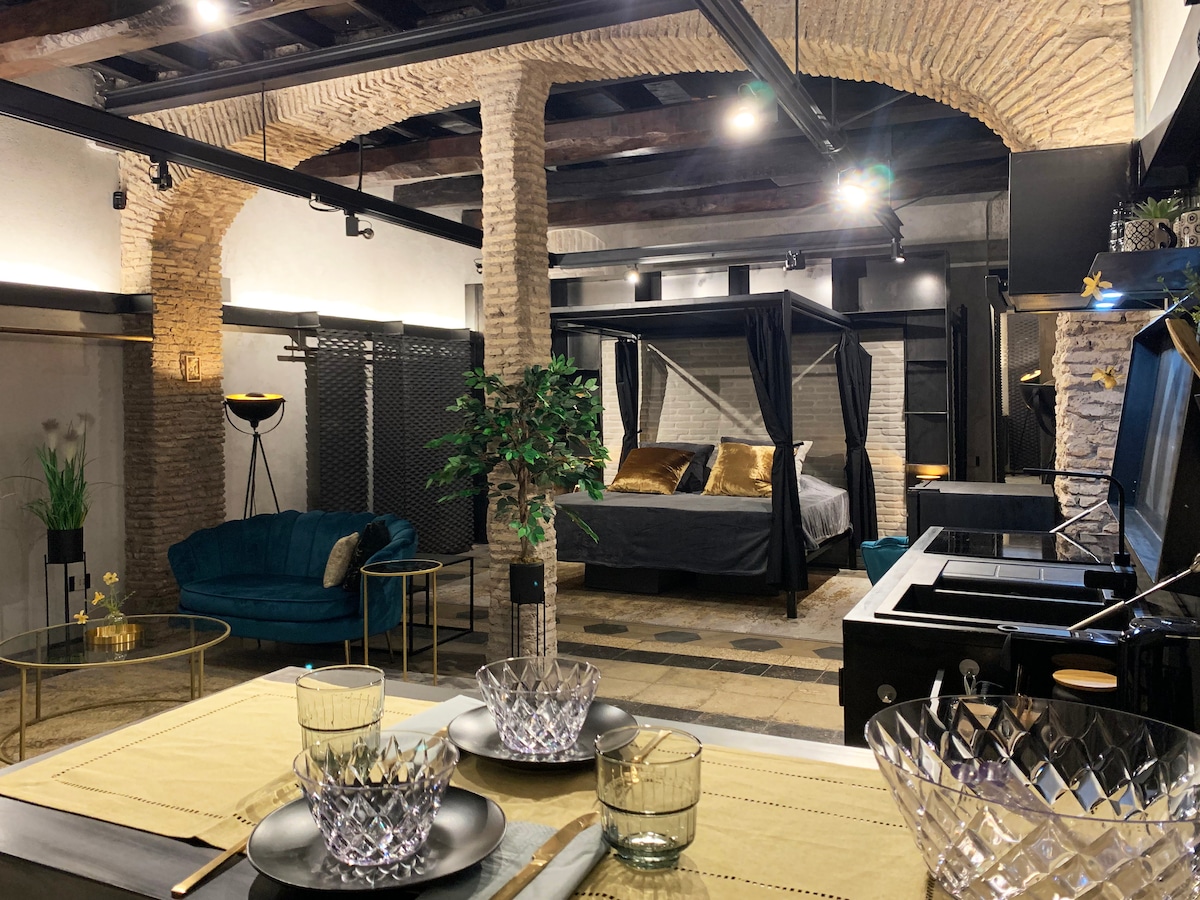An open living space features exposed stone columns and wooden beams overhead. A four-poster bed is positioned in the background, complemented by gold accents. A glass dining table is set with elegant dishware, while a plush couch adds comfort to the inviting area.