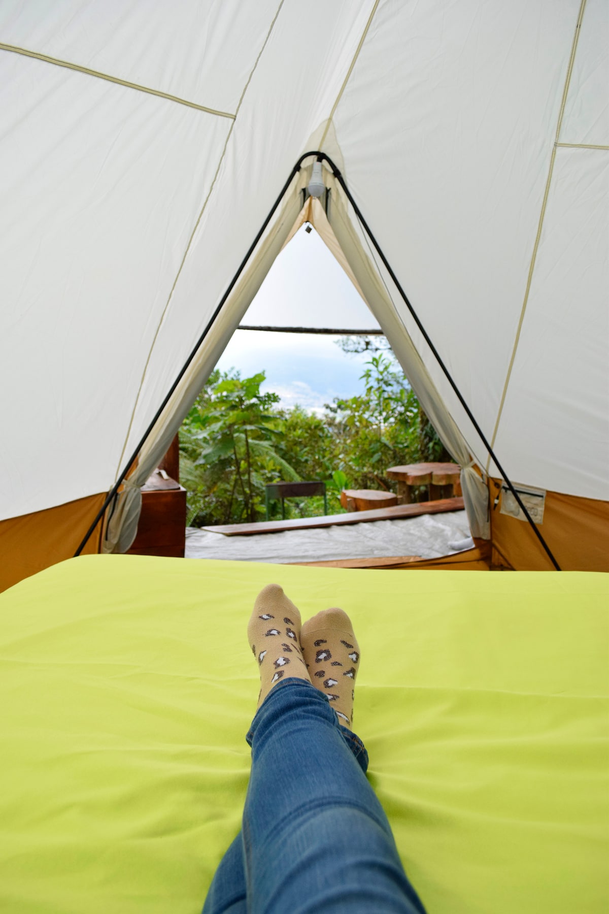 Inside the glamping tent, a spacious bed is adorned with a bright green coverlet. A view of the outdoor area is visible through the entrance, featuring natural greenery and a wooden table. A person’s feet, wearing patterned socks, are positioned comfortably on the bed.