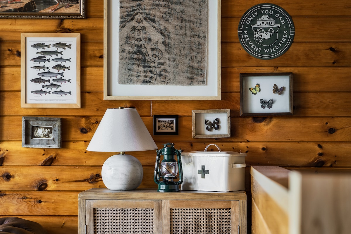 A rustic wall features a variety of framed artwork, including fish and butterflies, displayed alongside wooden paneling. A contemporary lamp and a vintage lantern are placed on a wooden table, enhancing the cabin's homey aesthetic.