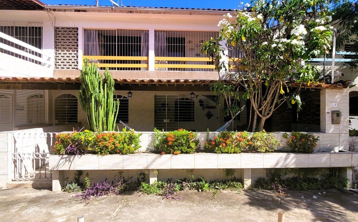 The front of a single-story house is framed by a lush garden featuring colorful flowers and green plants. A shaded porch is visible, complemented by a railing and tile roof. The structure is divided into sections, with large windows allowing natural light to enter.