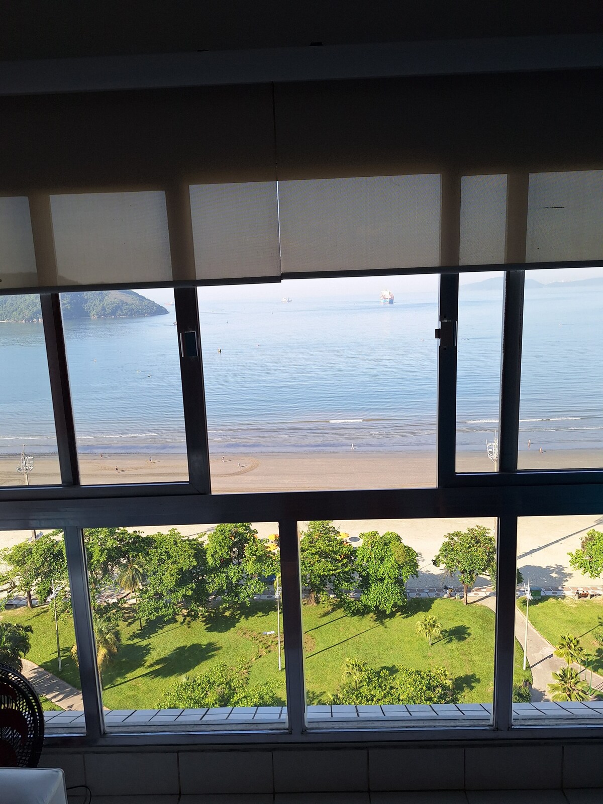 A clear view of the ocean is framed by large windows, revealing a sandy beach and gentle waves. Lush greenery is visible along the shoreline, creating a serene coastal landscape. The soft light enhances the tranquil atmosphere of this seaside location.