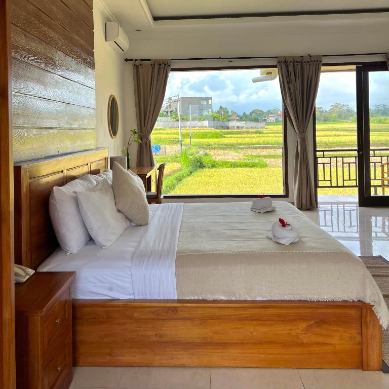 A spacious room is highlighted by a wooden bed and soft bedding, with large windows offering views of lush rice fields. Natural light fills the space, complemented by soft curtains. Two folded towels are neatly placed on the bed, enhancing the serene atmosphere.