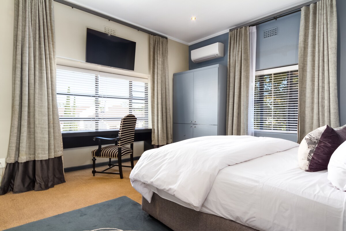 A spacious bedroom is presented with a large, comfortable bed adorned with white linens. A flat-screen TV is mounted on the wall, and natural light enters through the window framed by elegant curtains. A stylish chair sits next to a built-in cabinetry.