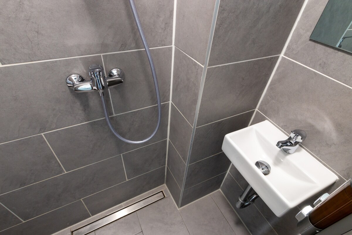 The updated bathroom presents sleek grey tiles and modern fixtures. A compact shower is equipped with a chrome showerhead, alongside a small sink mounted on the wall. A clear mirror above the sink completes the minimalist design.