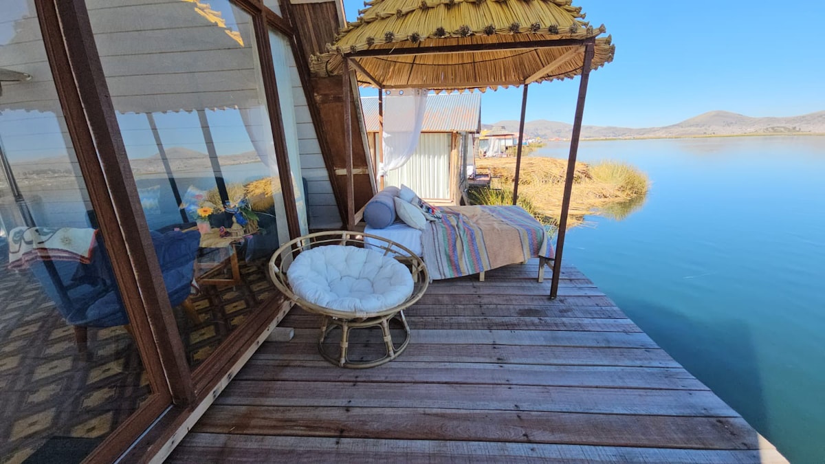 An outdoor seating area features a cozy bed adorned with a colorful blanket, surrounded by nature. A thatched-roof gazebo stands nearby, offering shade. The calm waters of a lake reflect the clear sky, while distant mountains create a serene backdrop.