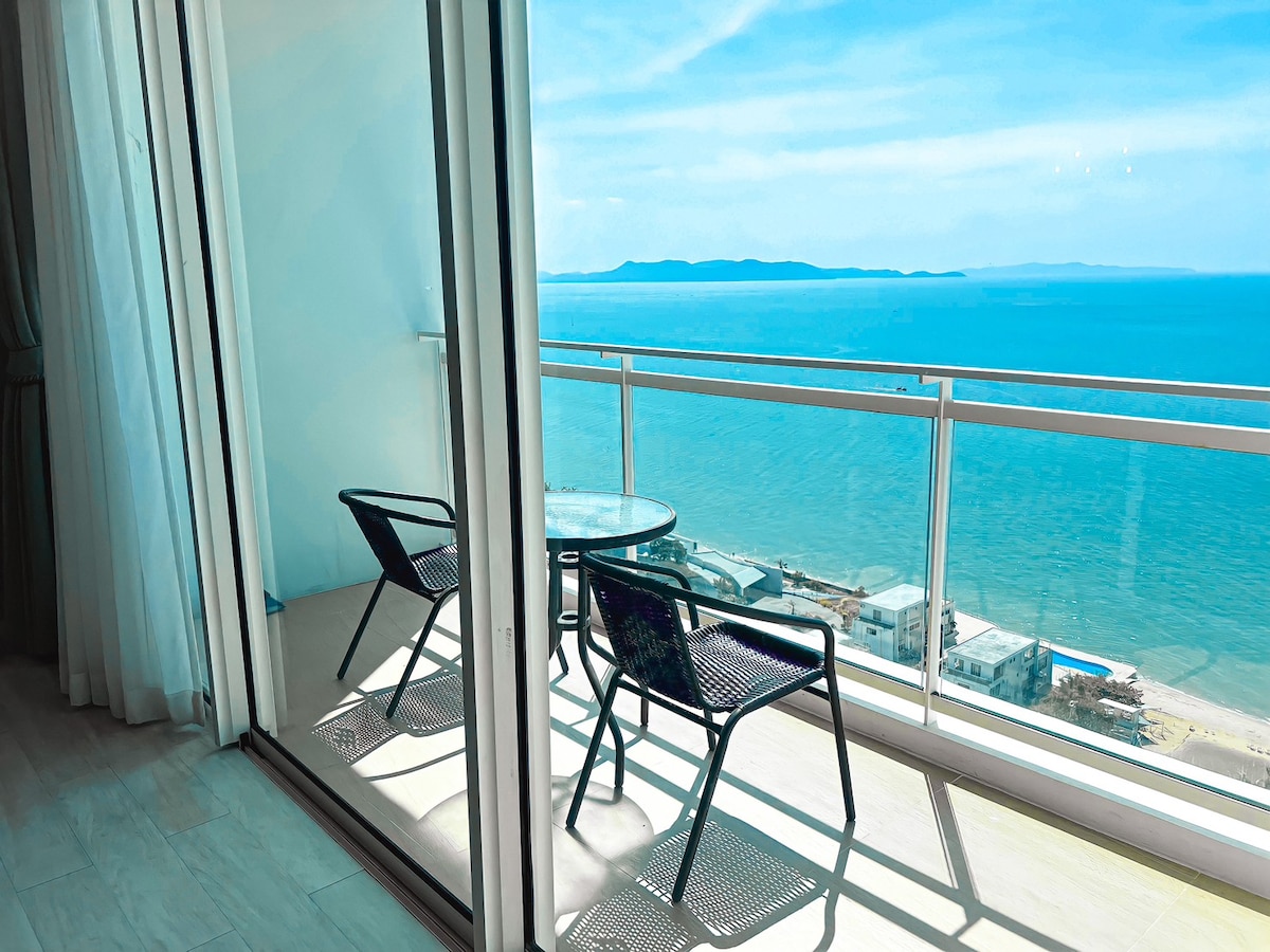 A private balcony showcases breathtaking sea views through expansive sliding glass doors. Two black wicker chairs and a small round table are positioned on the light-colored tiled flooring, providing a serene space to enjoy the coastal scenery.