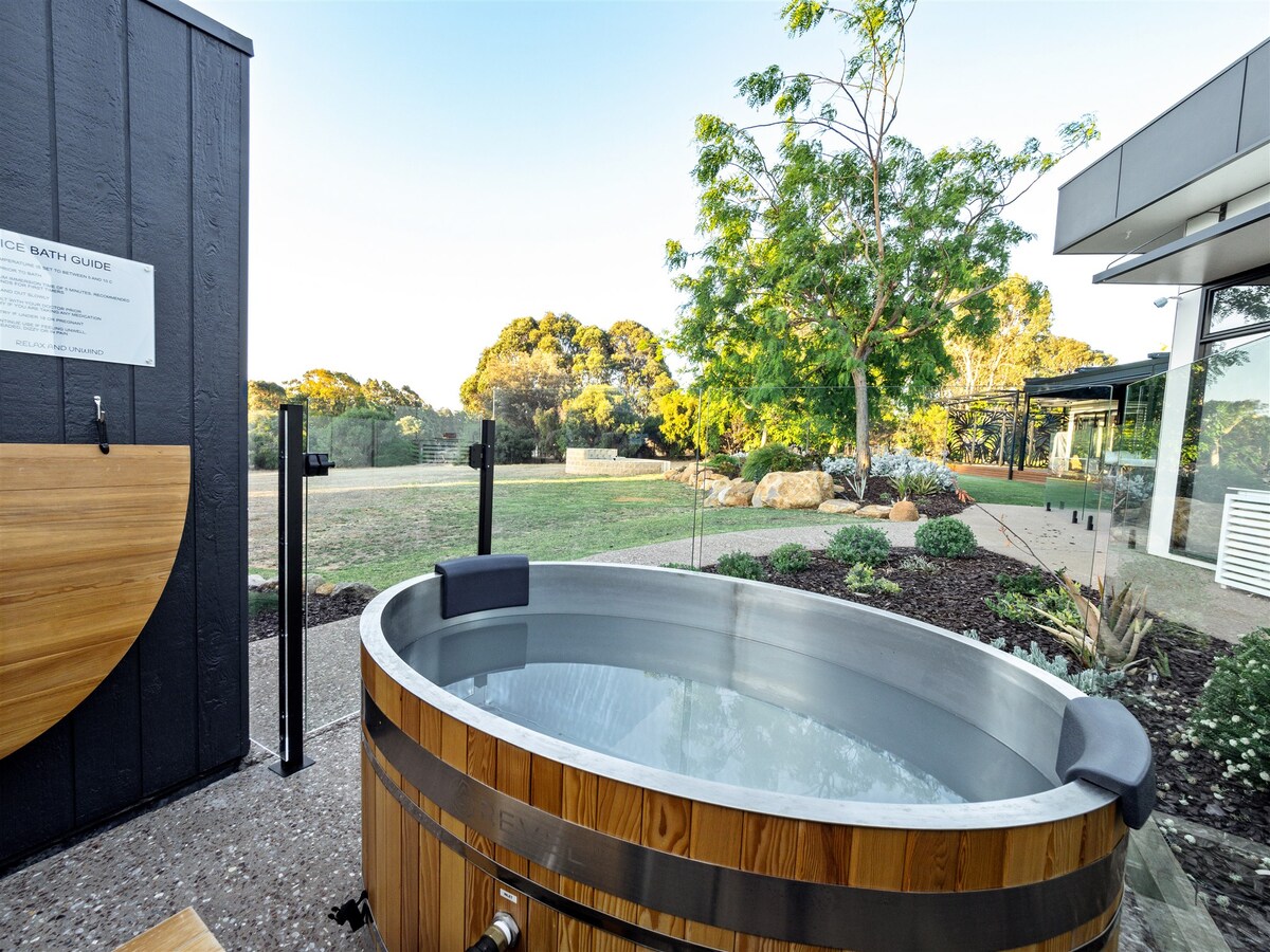 An outdoor ice bath is set against a backdrop of lush greenery and manicured gardens. The wooden tub features a sleek metallic edge and is surrounded by natural landscaping, inviting relaxation within a serene environment.