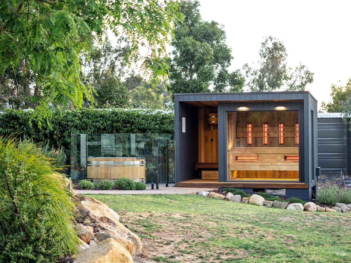 An outdoor wellness area features a modern infrared sauna with glowing red panels. Adjacent is a wooden ice bath enclosed by glass, providing an inviting space for relaxation. Lush greenery surrounds the area, enhancing the serene environment.