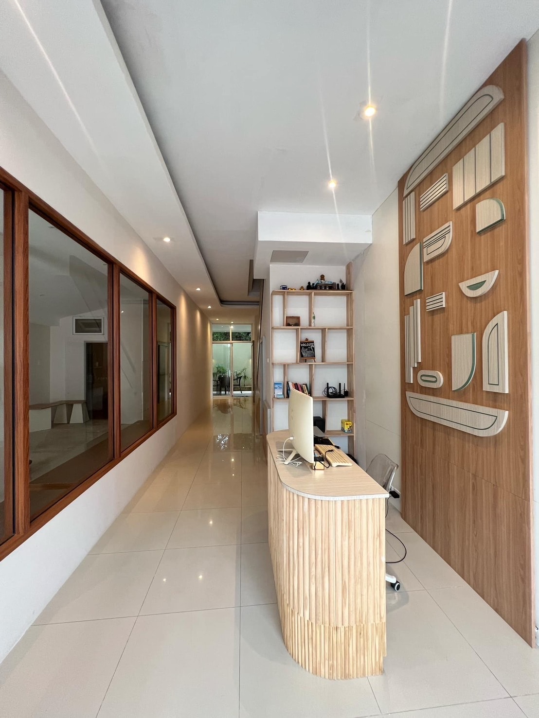 A modern reception area is highlighted by natural wood elements and sleek design. The space features a curved wooden desk, minimal furniture, and large windows allowing ample natural light. A decorative wall design adds texture, contributing to the overall contemporary aesthetic.