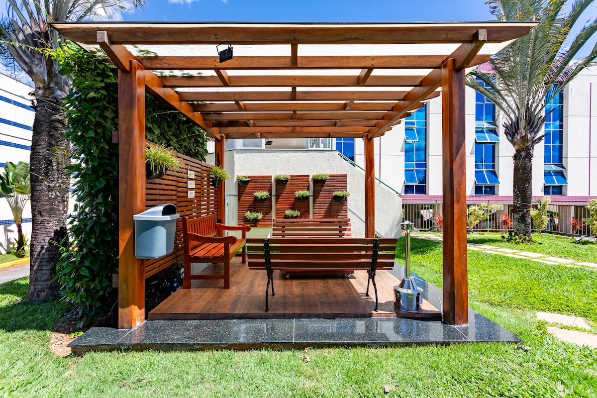 An outdoor seating area is framed by a wooden structure. It features a wooden bench and a red chair, surrounded by greenery and decorative plants. The flooring is wooden, and a clean pathway is visible, leading to a well-maintained lawn.