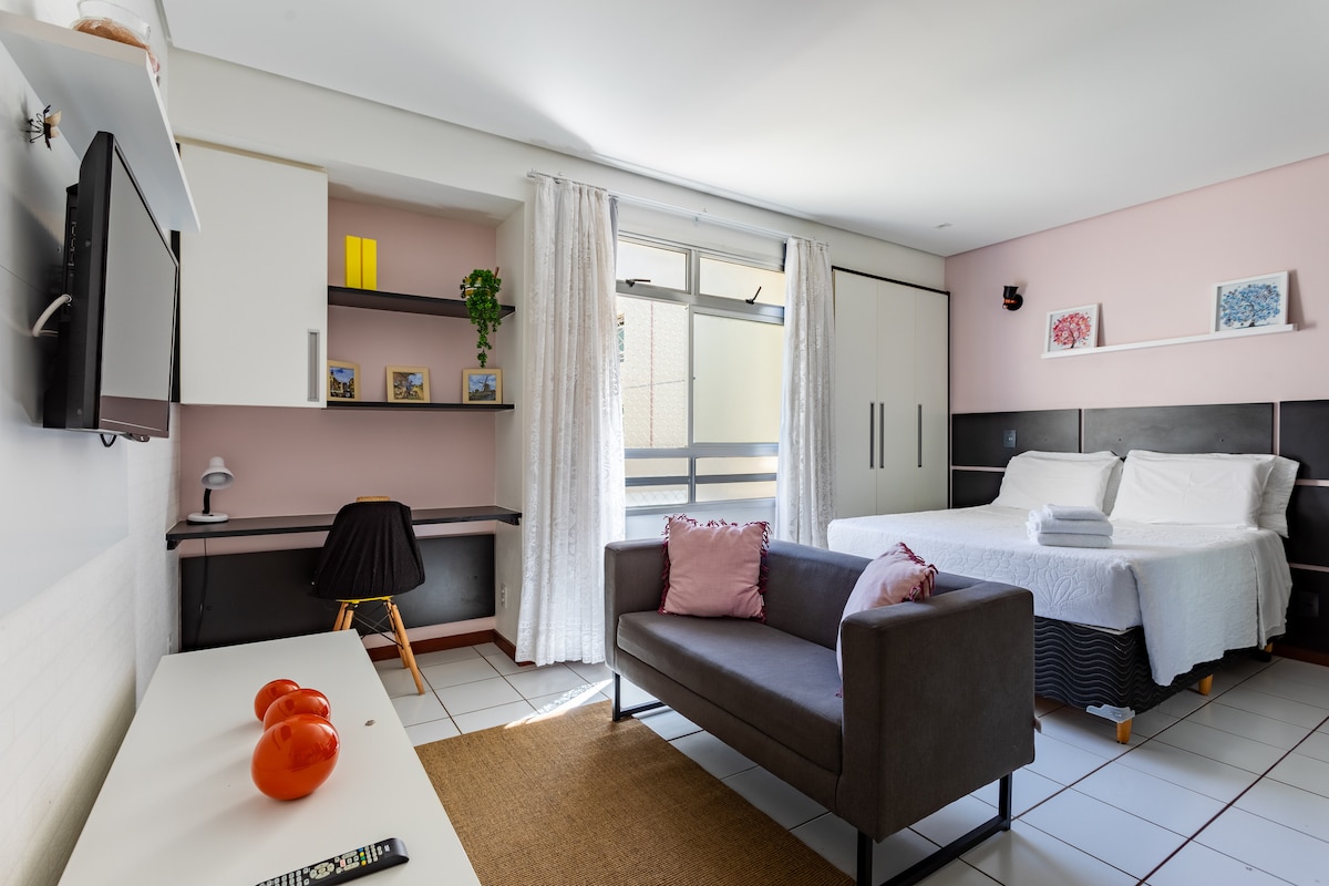 A well-organized studio space is presented, featuring a comfortable bed covered with fresh linens. A cozy sofa is positioned nearby, and a desk area is equipped with a lamp. Natural light enters through large windows, illuminating the pastel wall accents.