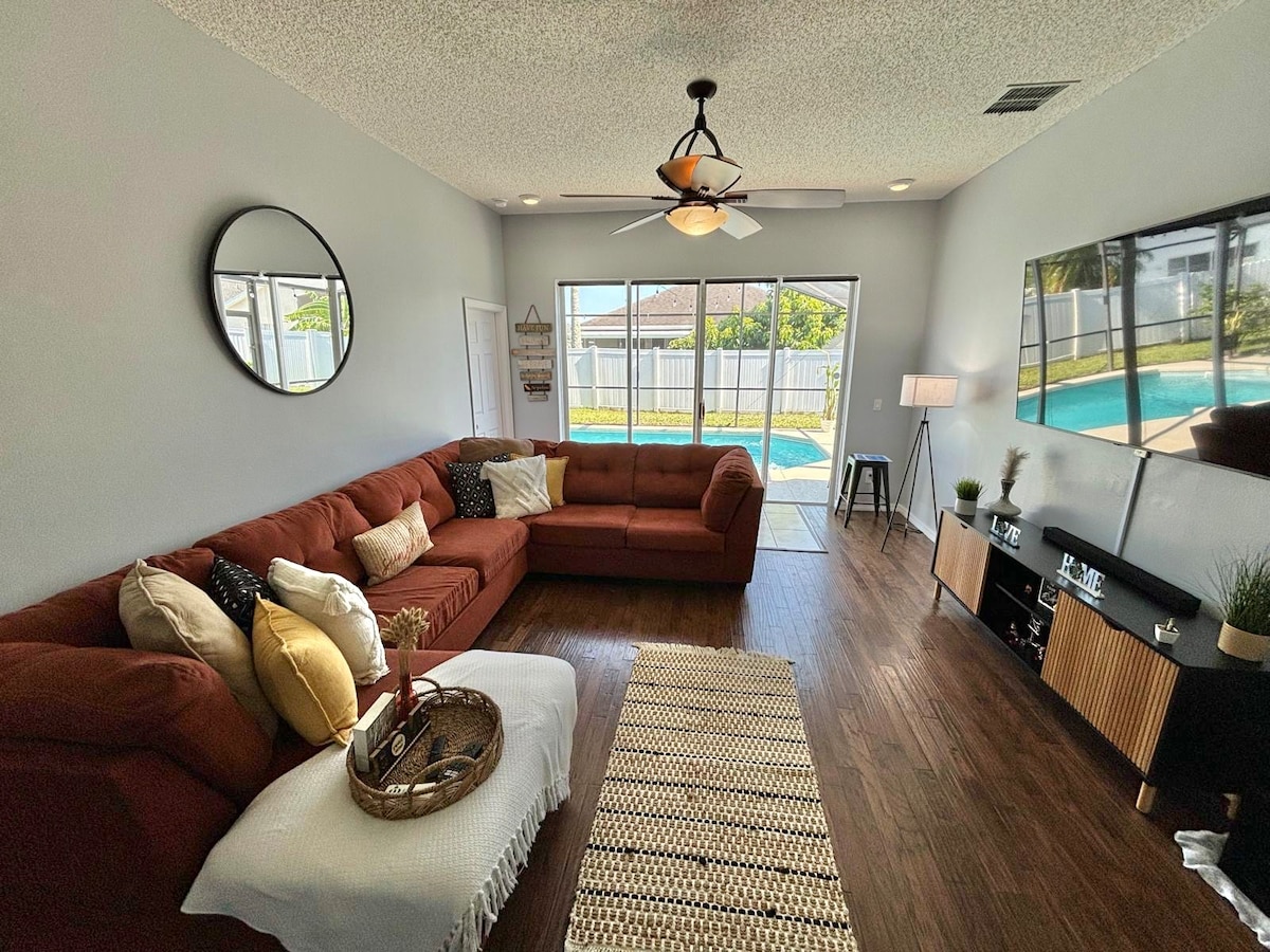 A spacious living area is depicted, featuring a large sectional sofa in earthy tones, adorned with decorative pillows. A soft area rug lies beneath. A sliding glass door opens to a pool area, framed by natural light. A media console and lamp complete the inviting space.