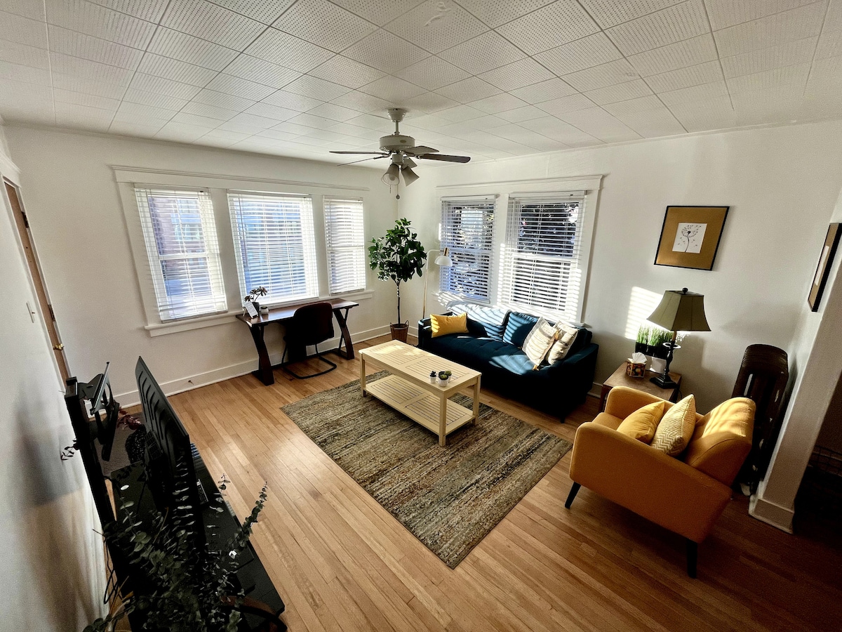 The living area features natural light streaming through multiple windows. A dark blue sofa is paired with a mustard-yellow armchair, and a wooden coffee table sits on a textured area rug. A desk is positioned near the windows, complemented by potted plants.