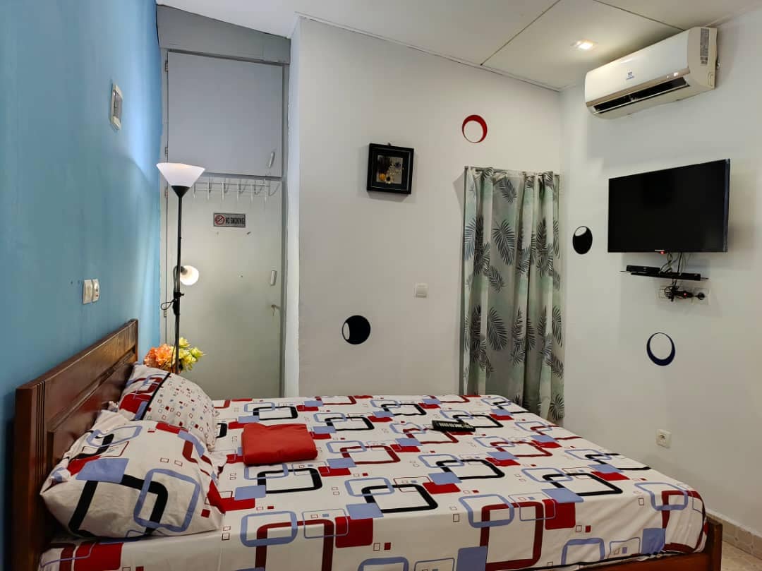 A cozy studio bedroom is adorned with a patterned bedspread featuring geometric shapes. A compact television is mounted on the wall, and a decorative curtain partially covers a window. Warm light is provided by a floor lamp, creating a cheerful ambiance.