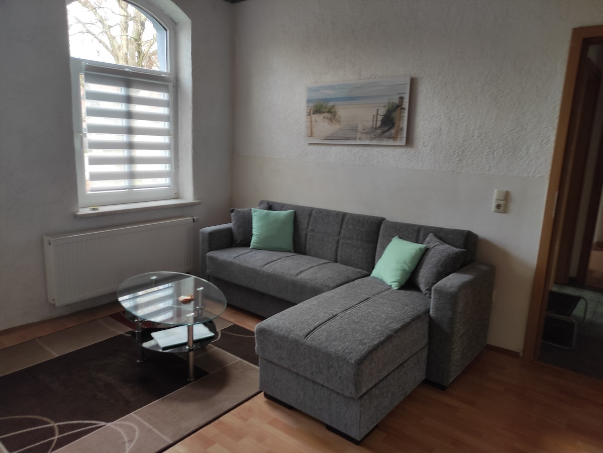 A cozy living area features a gray sectional sofa with two light green cushions, positioned beside a circular glass coffee table. Natural light enters through a window with stylish horizontal blinds, illuminating the soft texture of the carpet on the wooden floor.