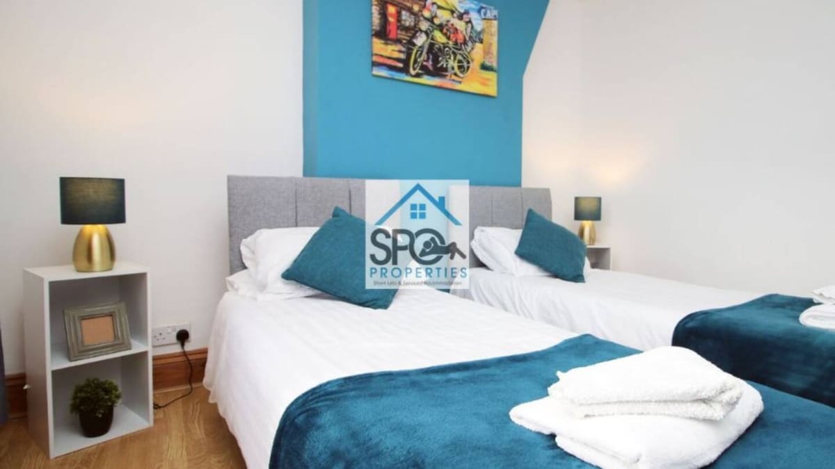 A well-appointed bedroom features two spacious beds with crisp white linens, accented by a teal throw blanket and decorative pillows. Modern lamps illuminate the space, and a small plant enhances the fresh look. The walls are painted a bright white, providing a clean backdrop.