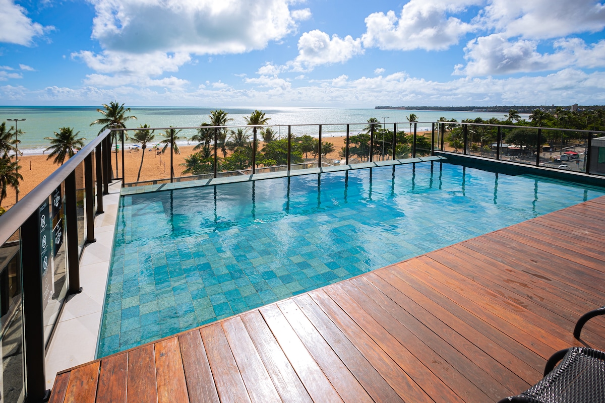 An inviting rooftop pool offers uninterrupted views of the ocean and sandy beach below. Palm trees line the shoreline, and the clear blue sky is reflected in the water, creating a tranquil setting. A wooden deck borders the pool, providing a space for relaxation.