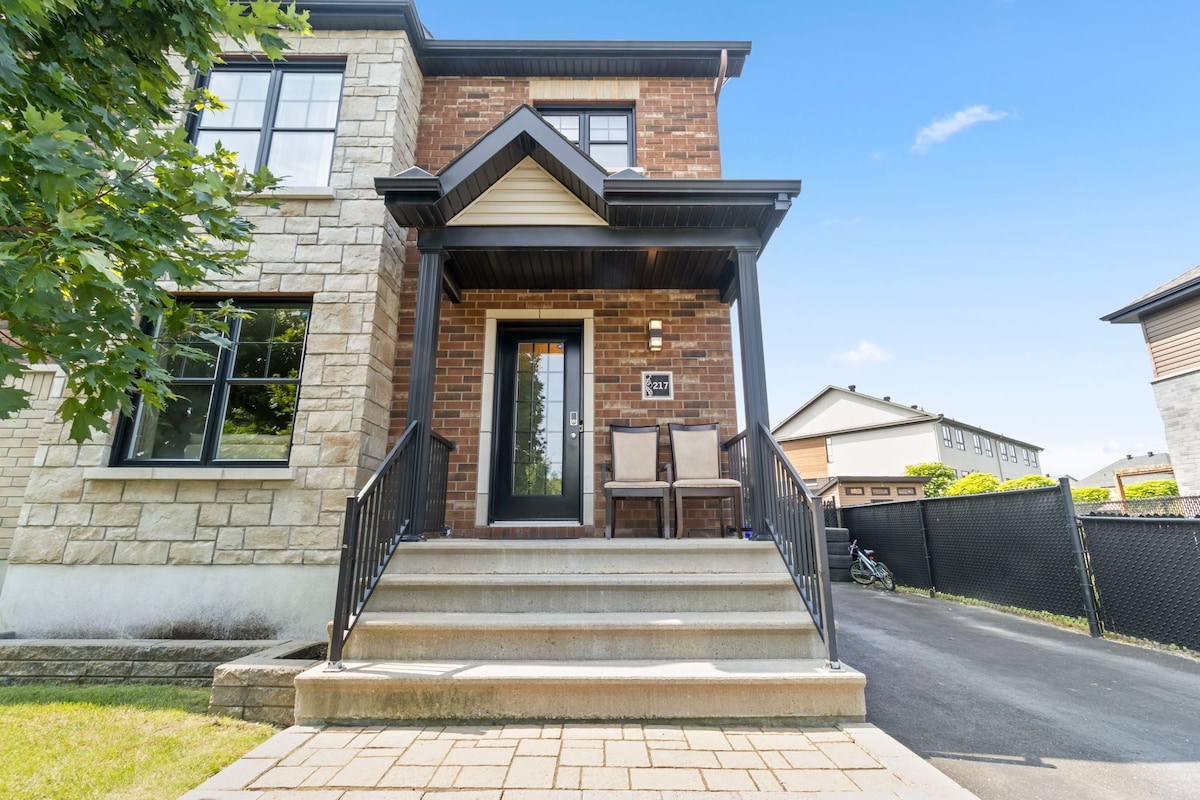 A welcoming entrance is highlighted by a set of steps leading to a front door, flanked by two outdoor chairs. The house features a blend of stone and brick exteriors, with large windows allowing natural light to enter.