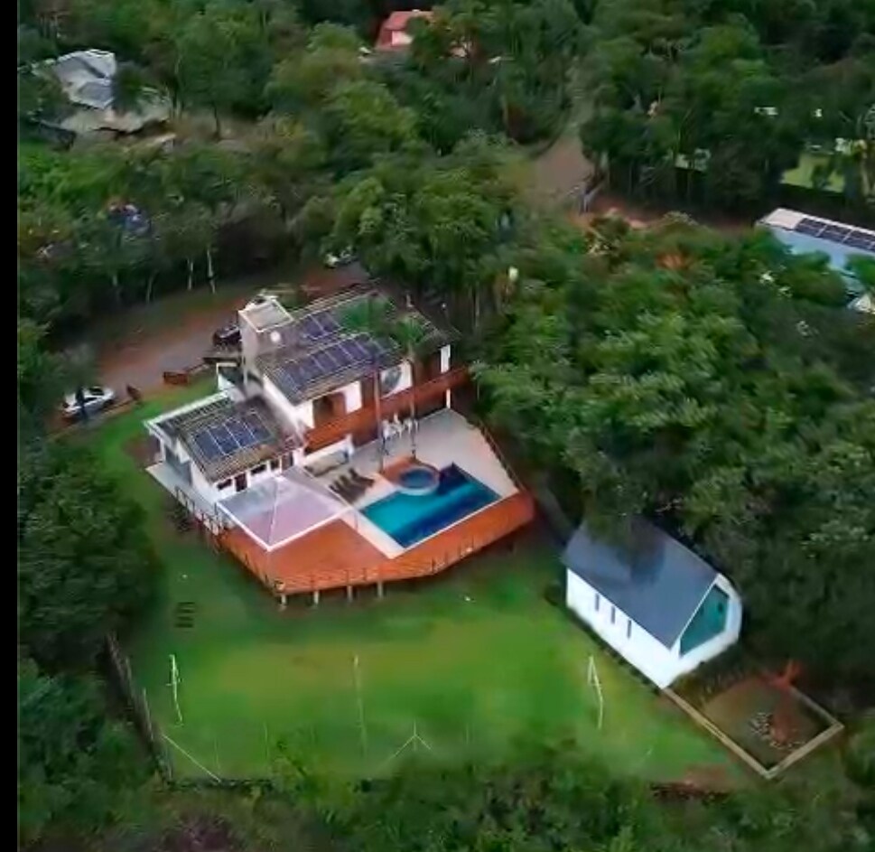 An aerial view captures a modern home nestled within lush greenery. A large pool is bordered by a spacious deck, and solar panels are installed on the roof. The surrounding landscape features trees and a small white chapel nearby.