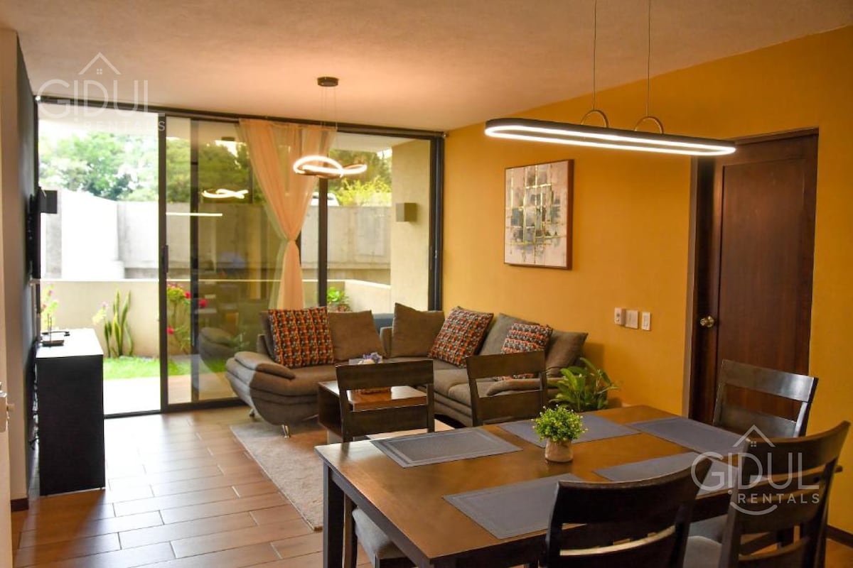 An open living and dining area showcases a modern design, featuring a spacious dining table with dark wood chairs. A comfortable sofa is arranged nearby, with colorful cushions adding accents. Large glass doors allow natural light to fill the space and provide a view of the garden outside.